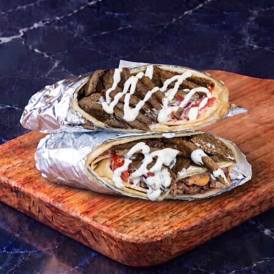 Two foil-wrapped gyros filled with seasoned meat, tomatoes, onions, and white sauce sit on a wooden serving board.