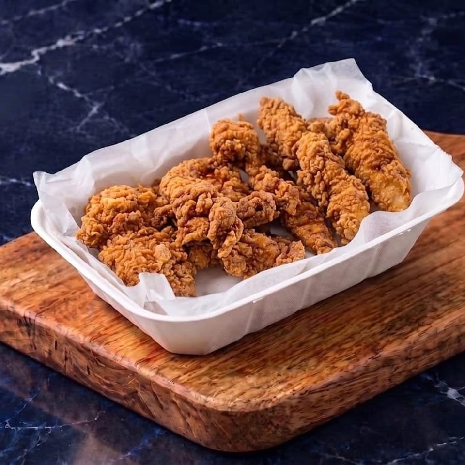Basket of crispy fried chicken strips on parchment, resting on a wooden serving board over a dark marble table.