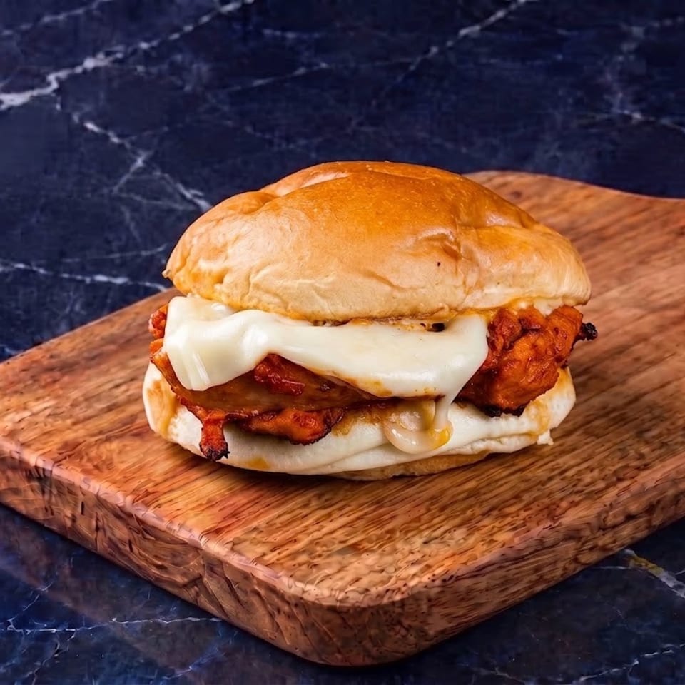 Loaded fried chicken sandwich with melted cheese and bacon on a wooden serving board against black marble.